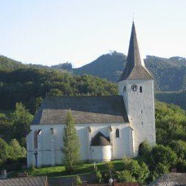Eine weiße Kirche mit Turm und Uhr liegt eingebettet in einem üppigen Tal, umgeben von Grün und Bergen.