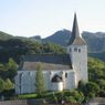 Eine weiße Kirche mit Turm und Uhr liegt eingebettet in einem üppigen Tal, umgeben von Grün und Bergen.