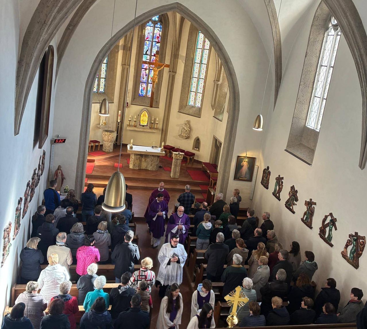 Eine große Gruppe von Menschen versammelt sich in einer Kirche. Priester stehen am Altar. Viele sitzen auf Kirchenbänken. Religiöse Symbole schmücken die Wände.