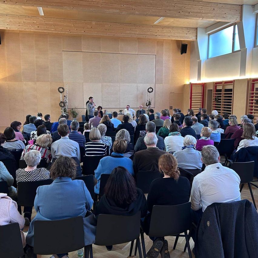 Ein Saal mit Holzwänden beherbergt eine Vorlesung. Mehrere Menschen sitzen in Stühlen vor einer Bühne mit Lautsprechern. Eine Person spricht auf der Bühne. Ein Kranz hängt an der Wand.
