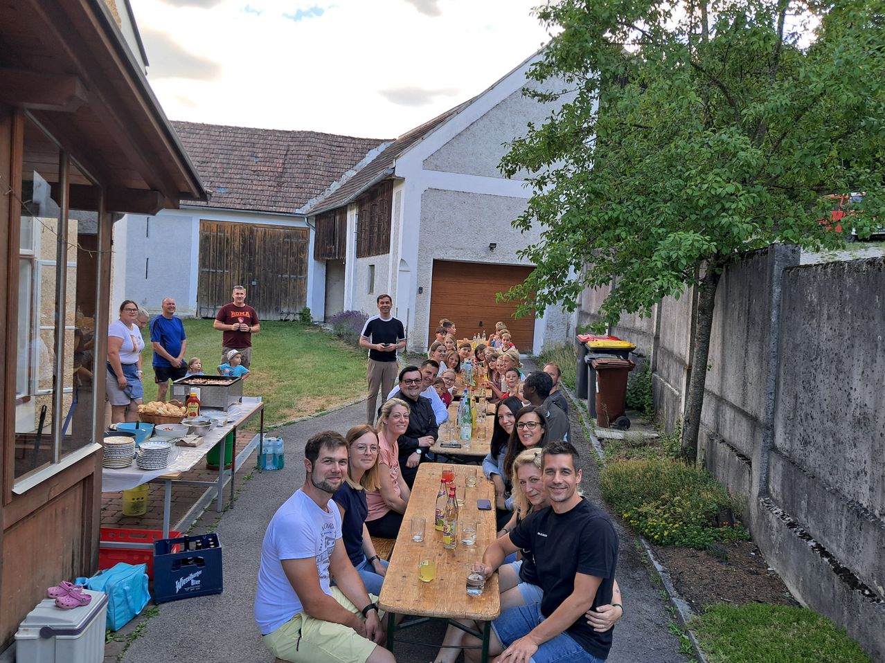 Eine Gruppe von Menschen sitzt um einen langen Tisch in einem Hinterhof, mit einem Haus und Bäumen im Hintergrund.