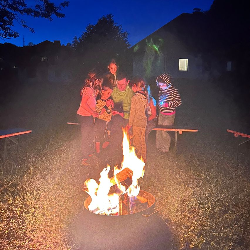 Eine Gruppe von Menschen, einschließlich Kindern, versammelt sich um ein Lagerfeuer bei Nacht, mit einem Haus und Bäumen im Hintergrund.