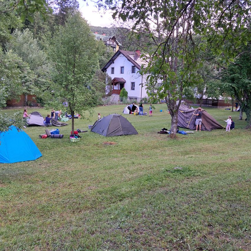 Eine Gruppe von Menschen zeltet auf einem Rasen mit mehreren aufgestellten Zelten, einige unter Bäumen. Ein Haus ist im Hintergrund sichtbar.