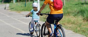 Ein Mann und ein Mädchen fahren auf einem Weg mit Fahrrädern. Das Mädchen trägt einen Helm und der Mann hat einen Rucksack. Sie tragen beide Turnschuhe. Das Fahrrad des Mannes hat eine Klingel und einen Korb.
