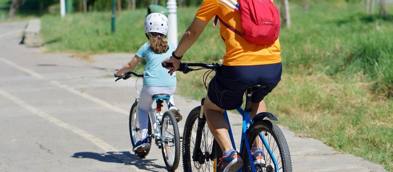 Ein Mann und ein Mädchen fahren auf einem Weg mit Fahrrädern. Das Mädchen trägt einen Helm und der Mann hat einen Rucksack. Sie tragen beide Turnschuhe. Das Fahrrad des Mannes hat eine Klingel und einen Korb.