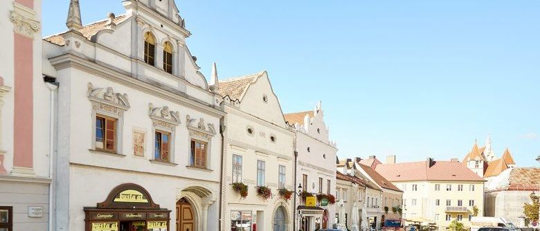 Eine Stadtstraße mit geparkten Autos, mit weißen Gebäuden mit verzierten Dekorationen und einem klaren blauen Himmel. Mehrere Geschäfte säumen die Straße, eines davon hat ein Schild, das 'Computer' besagt.