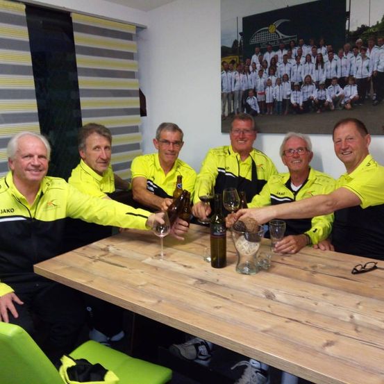 Sechs Männer in gelben Jacken stoßen mit ihren Gläsern und Flaschen an einem Holztisch an, mit einem Foto einer Tennismannschaft dahinter.