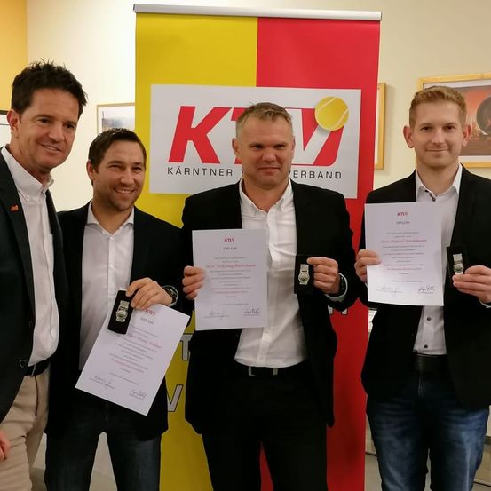 Vier Männer stehen vor einem gelben und roten Banner mit dem Logo und dem Text 'KTV'. Sie halten Urkunden und lächeln für ein Foto. Der Mann auf der linken Seite trägt ein weißes Hemd und Khakihosen.