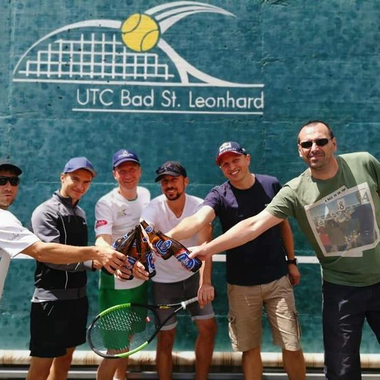 Fünf Männer stehen vor einem Tennisplatz. Sie tragen alle Mützen und Sonnenbrillen. Ein Mann hält eine Bierflasche. Ein anderer Mann hält einen Tennisschläger. Die Wand hinter ihnen hat ein Tennisball-Logo.