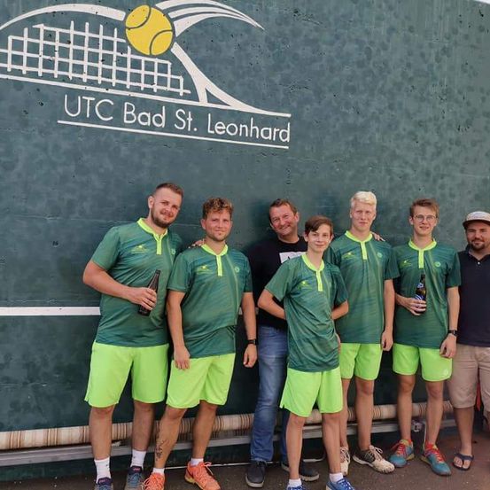 Eine Gruppe von Menschen in grünen Uniformen posiert für ein Foto vor einem Tennisplatz. Sie halten Bierflaschen und der Hintergrund zeigt das Logo von UTC Bad St. Leonhard.