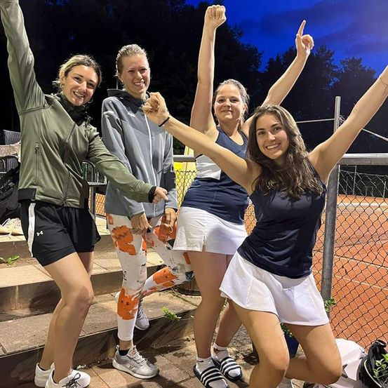 Fünf Frauen, wahrscheinlich Tennisspielerinnen, stehen auf einer Treppe und feiern. Sie tragen Tennisuniformen und Turnschuhe. Die Frauen posieren für ein Foto mit erhobenen Händen. Hinter ihnen steht ein Zaun, und ein Tennisplatz ist sichtbar.