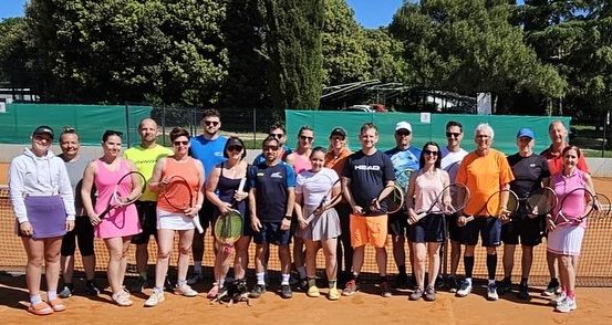 Eine Gruppe von Menschen steht auf einem Tennisplatz und hält Tennisschläger. Sie tragen Kleidung und Mützen in verschiedenen Farben. Der Platz ist von einem Zaun und Bäumen umgeben.