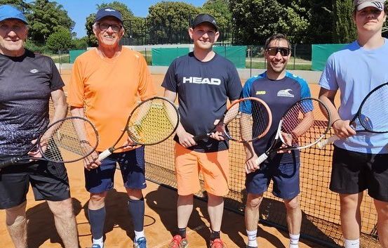 Fünf Männer stehen auf einem Tennisplatz und halten Schläger. Einer hält eine Tenniskugel. Sie tragen Freizeitkleidung. Einer trägt ein HEAD-Shirt. Der Hintergrund zeigt einen Zaun und Bäume.