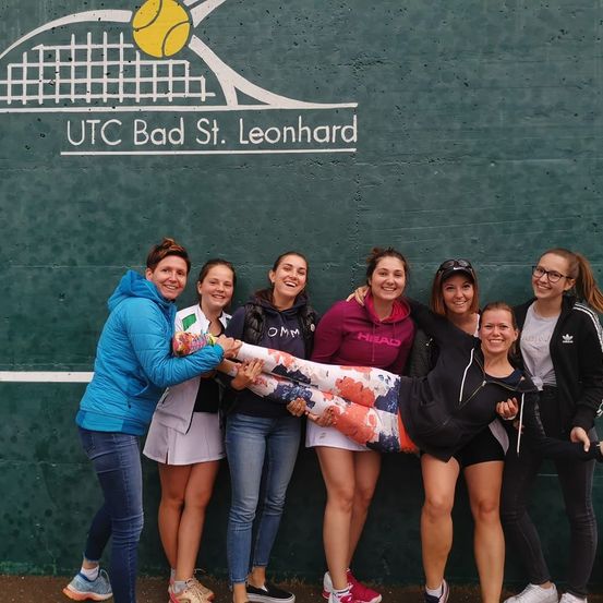 Eine Gruppe von Frauen posiert für ein Foto auf einem Tennisplatz. Sie stehen vor einer grünen Wand mit den Worten UTC Bad St. Leonhard.