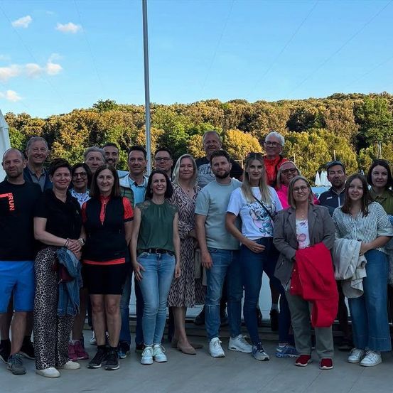 Eine Gruppe von Menschen steht lächelnd für ein Foto draußen zusammen. Vor ihnen befindet sich eine Kulisse aus Bäumen und blauem Himmel. Einige tragen Freizeitkleidung, andere Sportbekleidung.