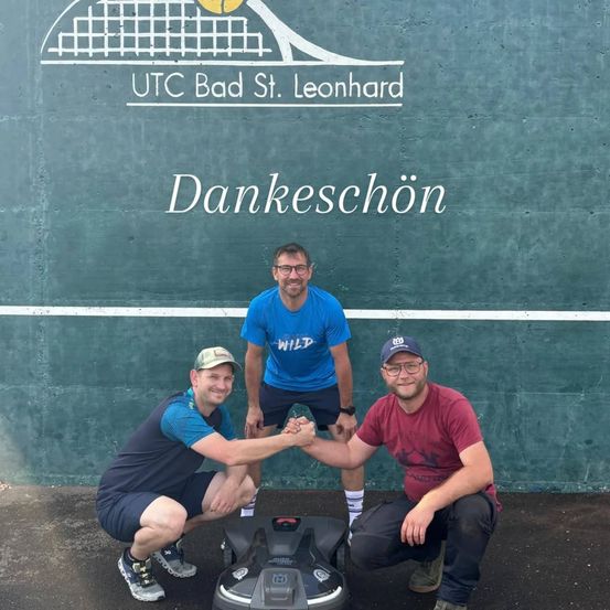 Drei Männer posieren vor einer Wand, lächeln und schütteln Hände. Ein Tennisplatzlogo ist an der Wand. Ein Mann trägt eine Brille, und die anderen beiden haben Turnschuhe an. Ein schwarzes Objekt steht vor ihnen.