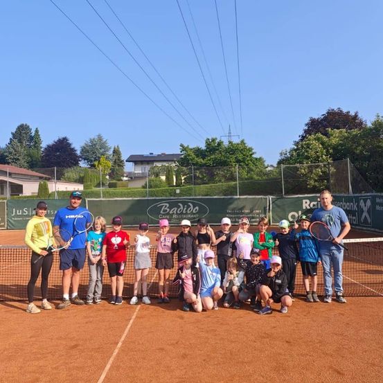 Eine Gruppe von Kindern und zwei Erwachsenen posiert für ein Foto auf einem Tennisplatz. Sie halten Tennisschläger und lächeln. Dahinter befindet sich ein Zaun mit Werbung.