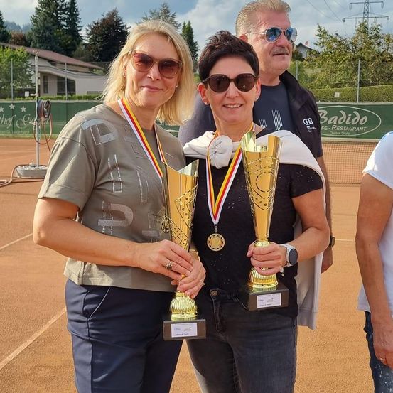 Zwei Frauen halten Pokale und stehen lächelnd auf einem Tennisplatz. Eine trägt eine Medaille, die andere hat eine Schärpe. Hinter ihnen beobachtet ein Mann. Im Hintergrund eine Mauer und Bäume.