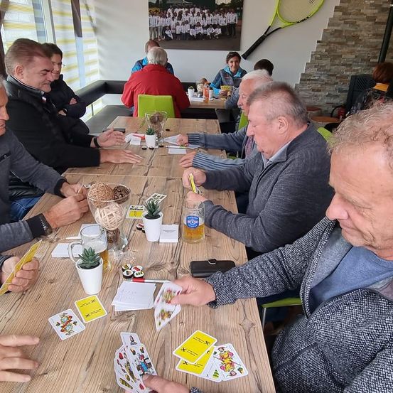 Eine Gruppe von Menschen sitzt um einen Tisch herum und spielt Karten, mit Getränken und Topfpflanzen. Eine Frau und ein Mann sitzen im Hintergrund an einem anderen Tisch.