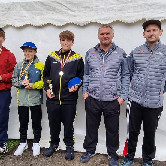 Fünf Personen stehen vor einem weißen Zelt, alle tragen Jacken und Medaillen. Der Mann in der Mitte hält einen blauen Gegenstand, möglicherweise ein Paddel. Zwei Jungen stehen auf beiden Seiten von ihm, jeder hält eine Trophäe. Sie scheinen einen Sieg zu feiern.