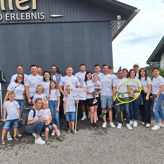 Eine Gruppe von Menschen in weißen Shirts steht vor einem Gebäude mit dem Wort 'Erlebnis' darüber. Sie posieren für ein Foto und halten Tennisschläger in den Händen.