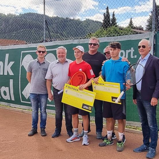 Eine Gruppe von Männern steht auf einem Tennisplatz und lächelt. Sie halten gelbe Schecks. Ein Banner mit Text steht hinter ihnen, und Berge sind in der Ferne.