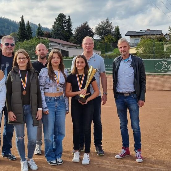 Eine Gruppe von Menschen steht auf einem Tennisplatz mit Medaillen um den Hals, eine hält eine Trophäe. Dahinter steht ein grüner Zaun mit einem Schild. Bäume und Häuser sind im Hintergrund.