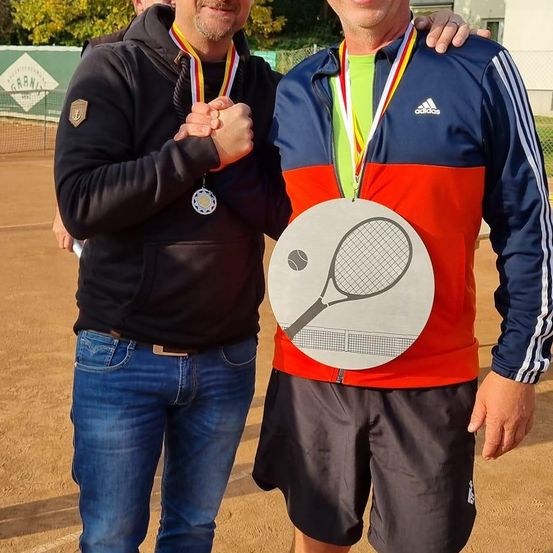 Zwei Männer, einer mit Medaillen, stehen auf einem Tennisplatz. Der Mann in Blau und Rot trägt eine große Medaille mit einem Tennisschlägerdesign.