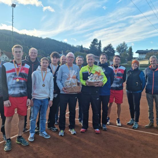 Eine Gruppe von Menschen mit Medaillen steht auf einem Tennisplatz und hält Geschenke. Ein Berg und Bäume sind im Hintergrund.