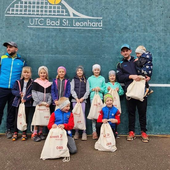 Gruppenfoto mit Kindern, Erwachsenen und einem Baby vor einer grünen Wand mit Tennisplatz-Logo und Name utc bad st. leonhard. Sie alle halten weiße Taschen mit Text und Logos.