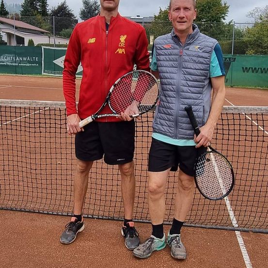 Zwei Männer stehen hinter einem Tennisnetz und halten beide Tennisschläger in der Hand. Einer trägt eine rote Jacke, der andere eine graue Weste. Sie lächeln für ein Foto auf einem Sandplatz.