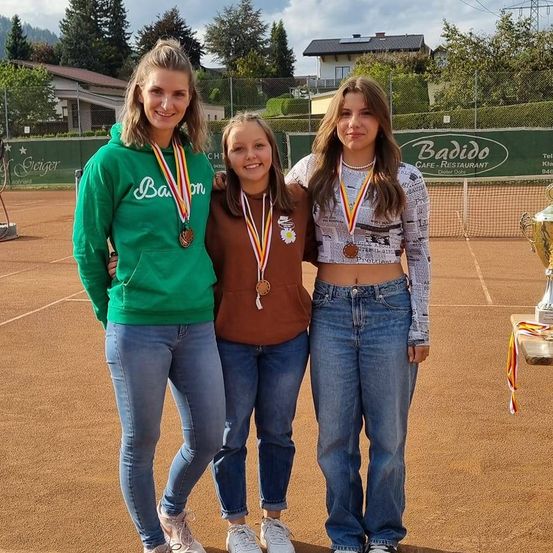 Drei Frauen stehen auf einem Tennisplatz und tragen Medaillen. Sie lächeln und posieren für ein Foto. Hinter ihnen befindet sich ein Zaun mit einem Schild, das 'Baddido Cafe-Restaurant' anzeigt. Bäume und Häuser sind im Hintergrund zu sehen.