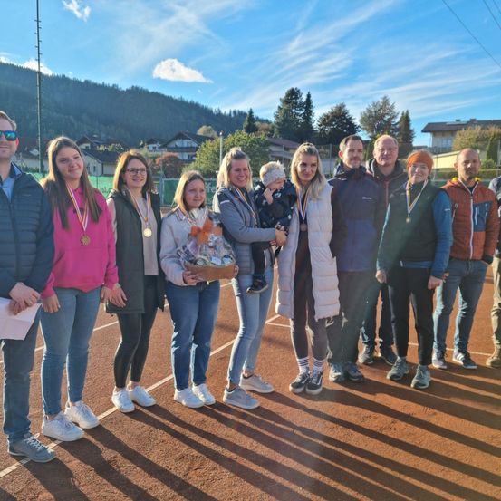 Eine Gruppe von Menschen steht auf einem Tennisplatz und trägt Medaillen. Eine Frau hält ein Baby und einen Geschenkkorb. Sie alle lächeln und posieren für ein Foto.