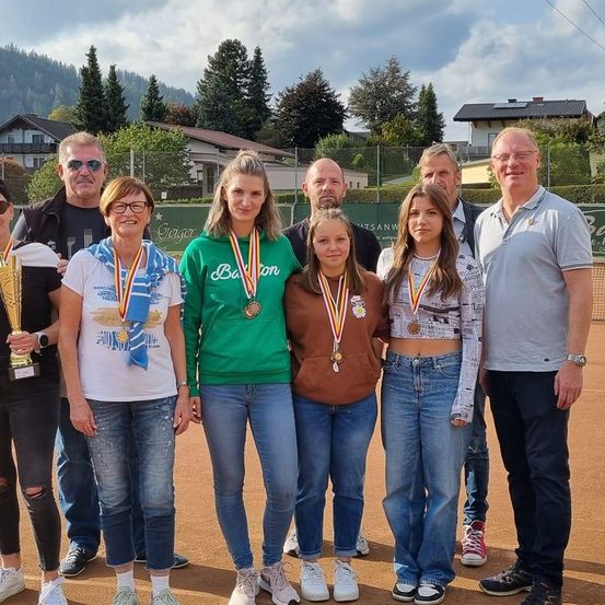 Mehrere Personen stehen auf einem Tennisplatz, einige halten Medaillen und Pokale. Sie sind lässig gekleidet, tragen Brillen, Sneakers und einige haben Schals. Hinter ihnen befinden sich Häuser, Bäume und ein Zaun. Der Himmel ist bewölkt.