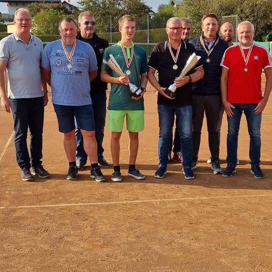 Eine Gruppe von Männern steht auf einem Tennisplatz, hält Trophäen, trägt Medaillen und lächelt für ein Foto.