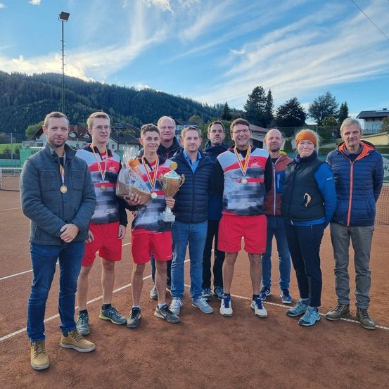 Eine Gruppe von Männern posiert auf einem Tennisplatz für ein Foto, mit Bergen im Hintergrund. Sie tragen Jacken und Sneaker, einer hält einen Pokal und einen Blumenstrauß.