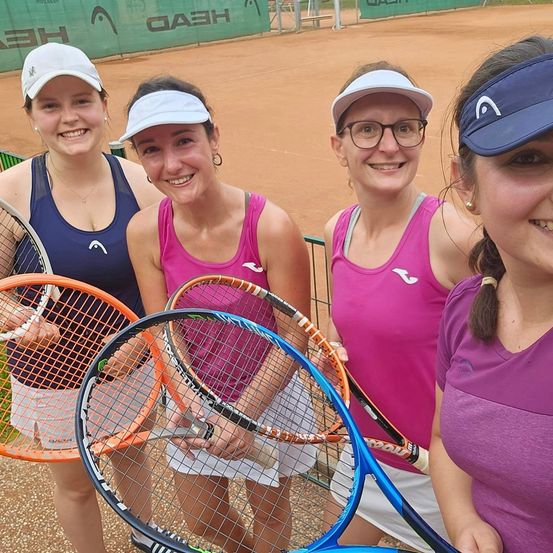 Vier Frauen stehen gemeinsam auf einem Tennisplatz, lächeln und halten Tennisschläger. Sie tragen passende rosa Sportoberteile und weiße Visiere. Der Platz ist eingezäunt.