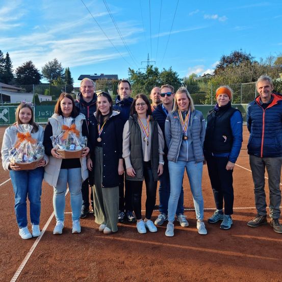 Eine Gruppe von Menschen steht auf einem Tennisplatz, trägt Medaillen und hält Geschenke. Der Himmel ist teilweise bewölkt. Einige tragen Brillen und Sneakers. Bäume und Häuser sind im Hintergrund sichtbar.