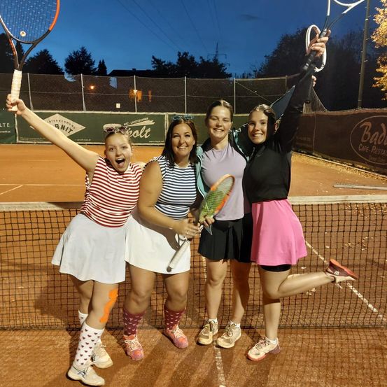 Vier Frauen posieren nachts auf einem Tennisplatz für ein Foto, alle halten Tennisschläger und tragen bunte Socken.