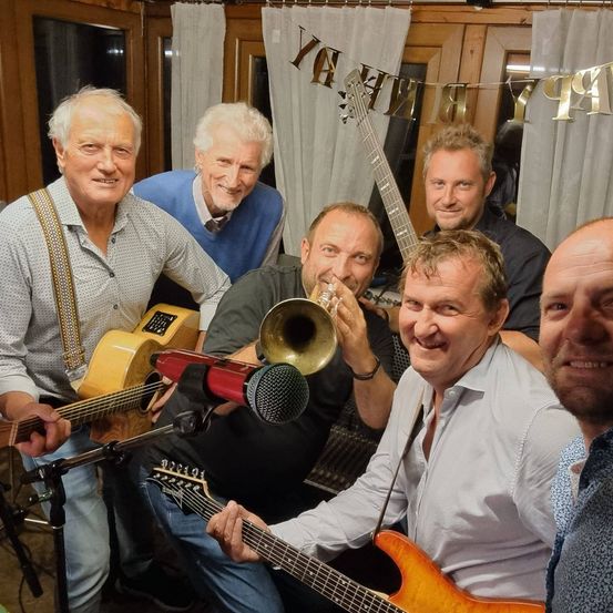 Fünf Männer lächeln und posieren für ein Foto. Einer spielt Trompete, ein anderer Gitarre. Sie sind von Musikinstrumenten umgeben. Ein Banner mit goldenen Zahlen 1979 im Hintergrund.