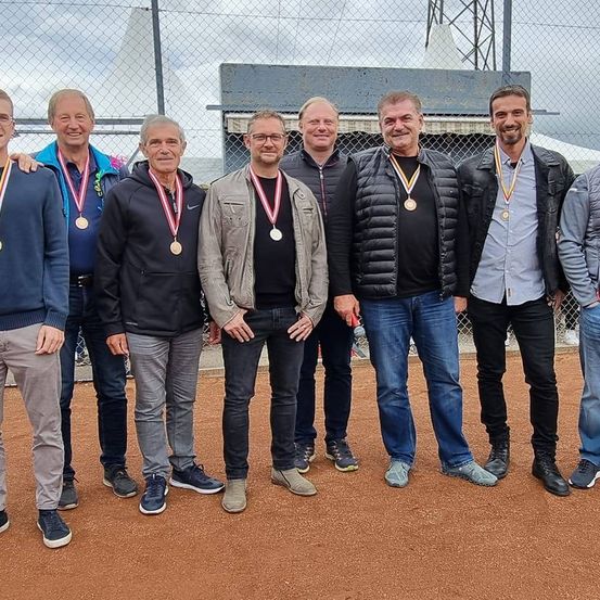 Sieben Männer stehen in einer Reihe und tragen Medaillen. Sie lächeln und posieren für ein Foto. Dahinter steht ein Zaun.