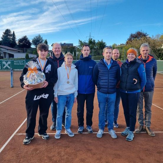 Eine Gruppe von Menschen auf einem Tennisplatz, zwei mit Medaillen, eine Frau hält einen Korb. Hinter ihnen ist ein Tennisnetz und ein Gebäude mit einem Schild. Bäume und ein Haus sind in der Ferne.