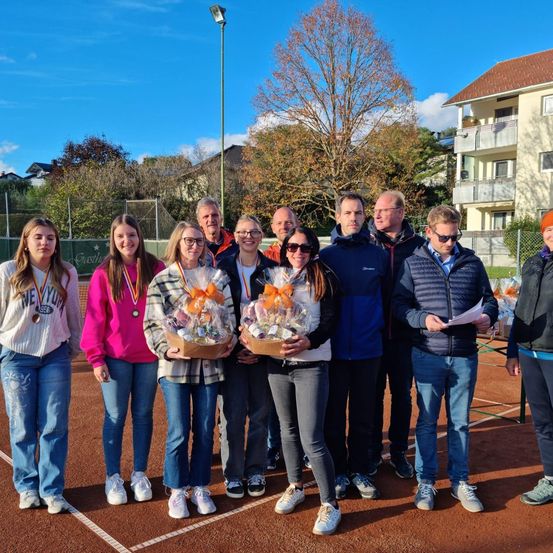 Eine Gruppe von Menschen steht auf einem Tennisplatz, hält Geschenke und steht unter einem blauen Himmel. Sie tragen Freizeitkleidung und einige haben Medaillen. Ein Gebäude mit Balkonen ist im Hintergrund.