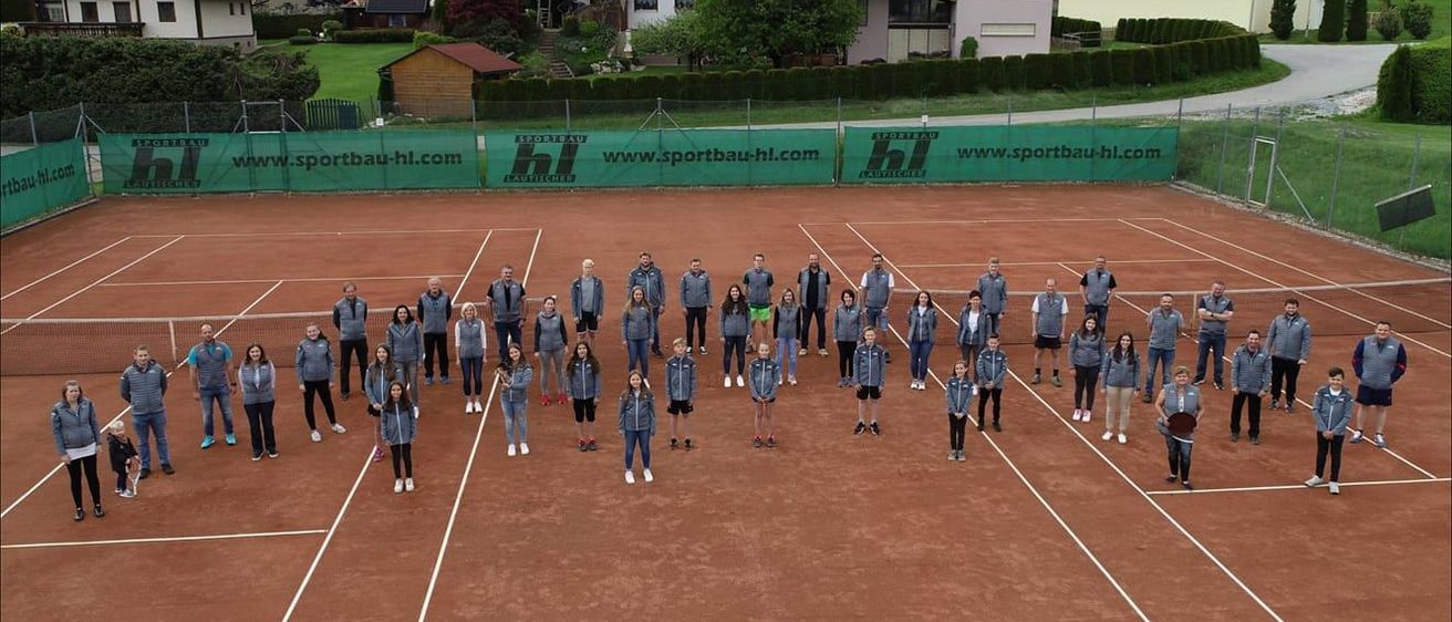 Eine Gruppe von Menschen in Jacken und Turnschuhen posiert für ein Foto auf einem Tennisplatz. Hinter ihnen ist ein grünes Banner mit der Website www.sportbau-hl.com.