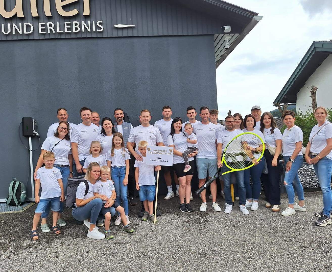 Gruppenfoto von Menschen mit Tennisschlägern vor einem Gebäude namens 'Erlebnis' mit bewölktem Himmel.