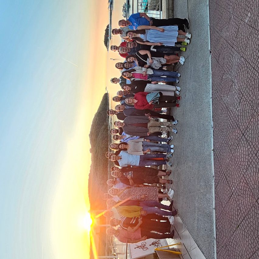 Eine Gruppe von Menschen steht in einer Reihe, mit Blick auf den Sonnenuntergang, mit Bergen und Meer im Hintergrund.