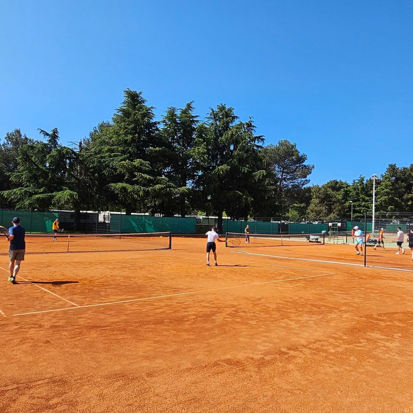 Mehrere Tennisspieler auf einem Sandplatz, umgeben von Bäumen, spielen an einem sonnigen Tag Tennis.