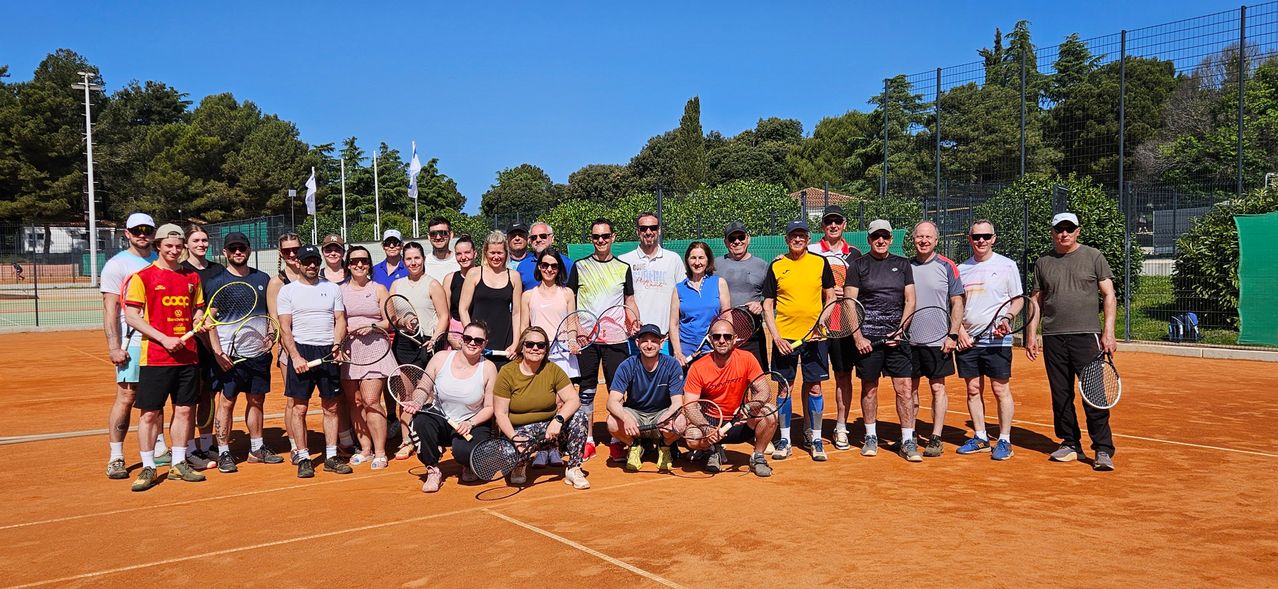 Eine Gruppe Tennisspieler in Freizeitkleidung posiert für ein Foto auf einem Tennisplatz mit Bäumen und einer Flagge im Hintergrund.