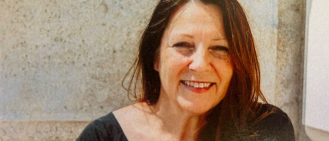 Eine lächelnde Frau mit kurzen braunen Haaren steht vor einer hellen Steinmauer und trägt ein schwarzes Oberteil.