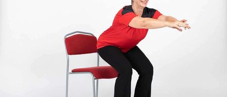 Eine lächelnde Frau in roter und schwarzer Kleidung sitzt auf einem roten Stuhl und streckt Arme und Beine, möglicherweise macht sie Yoga.